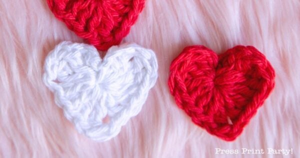 Three little crochet hearts—two red and one white—are arranged on a soft, pink, furry surface. The words "Press Print Party!" appear in the bottom right corner.
