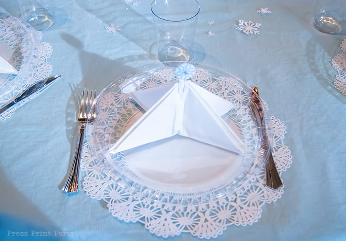 table setting with a tree napkin and a snowflake topper on a lace doily white For Christmas table decor ideas blue and silver winter wonderland decorations. Christmas tablescape for large event christmas party, diy holiday table setting. by Press Print Party!
