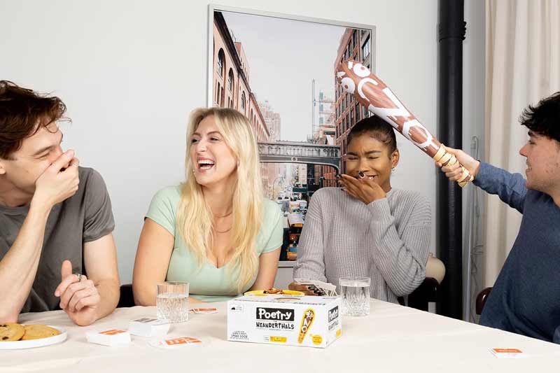 Four friends sit around a table, laughing like it's family game night. One playfully holds a large inflatable club over anotherβs head. Board game pieces and snacks cover the table, while a cityscape photo hangs on the wall.