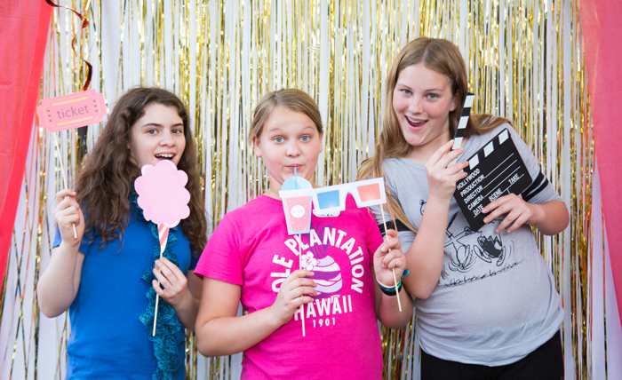 movie party photo booth with fun props. 3 girls having fun with props at a movie night party. Gold shimmering background and red curtains