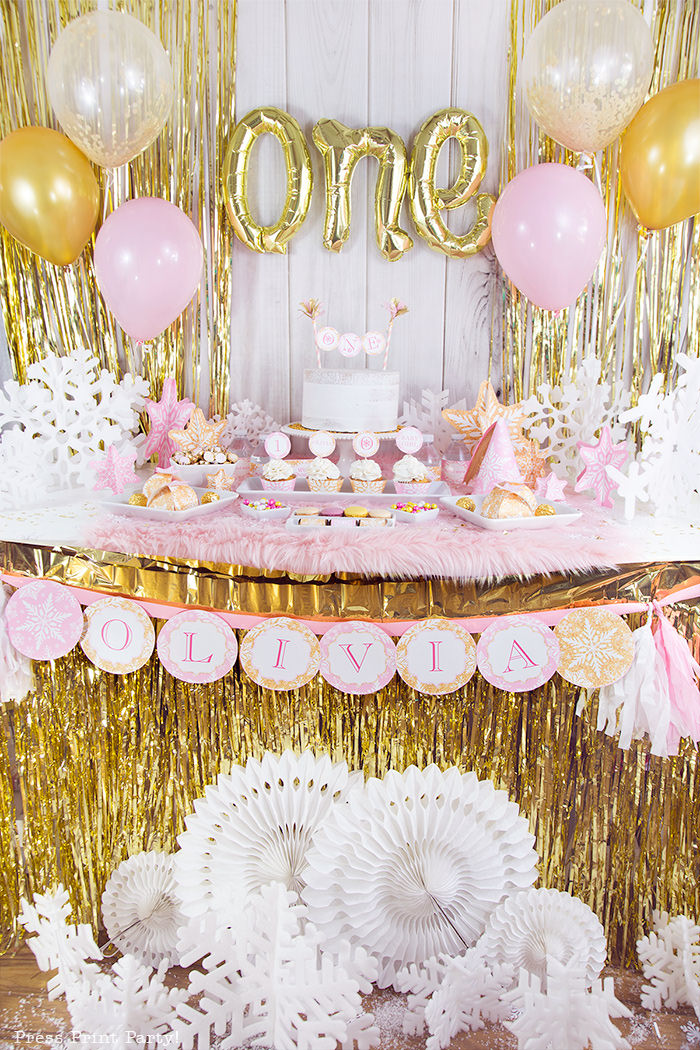 winter onderland first birthday girl party pink and gold decorations dessert table with onederland cake and cupcakes, printable snowflakes, favor boxes and chocolates, one inflatable balloons, pink and gold balloons and gold fringe backdrop - Press Print Party!