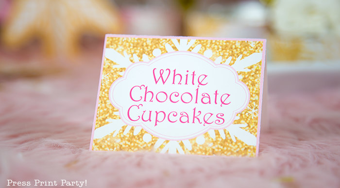 snowflakes place card, gold with pink lettering saying white chocolae cupcakes winter onederland gold and pink snowflakes kisses with printable labels winter onderland first birthday girl party pink and gold dessert table with onederland cake and cupcakes, printable snowflakes, favor boxes and chocolate - Press Print Party!