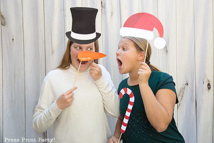 2 girls with Christmas photo booth props printables, one with a snowman hat and carrot nose, the other with santa hat and candy cane - Press Print Party!