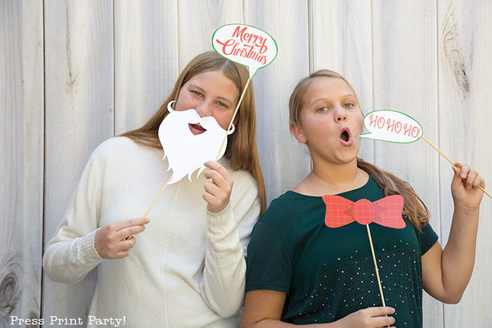 2 girls with Christmas photo booth props, one with santa beard and bubble saying Merry Christmas, the other with a printable bow and a bubble saying hohoho - Press Print Party!