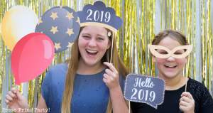 2 girls having fun with new year's eve photo booth props. 2019 tiara and speech bubble, balloons and mask. Press Print Party!
