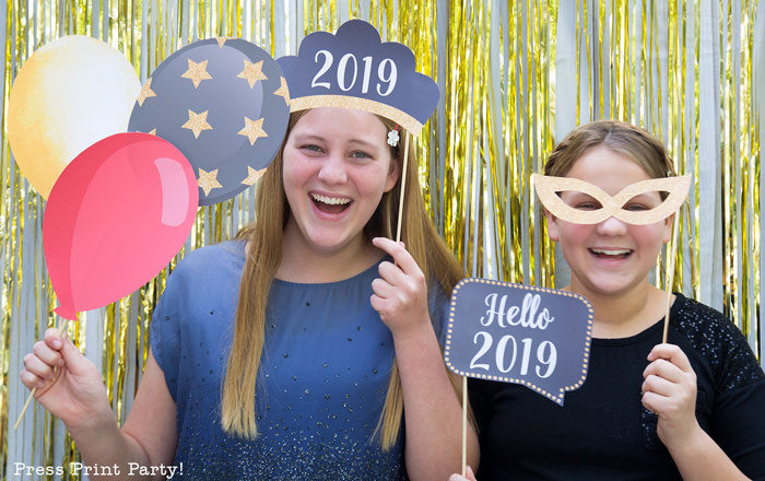 2 girls having fun with new year's eve photo booth props. 2019 tiara and speech bubble, balloons and mask. Press Print Party!