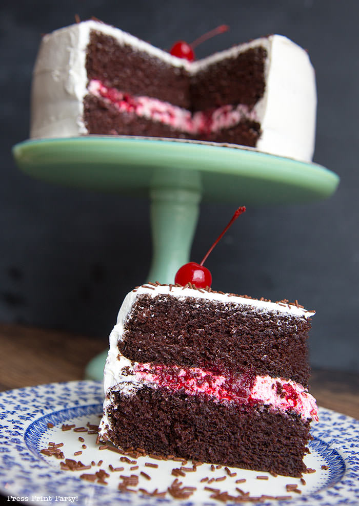 Moist chocolate cake with white frosting and berry filling on a green pedestal with a cherry on top and a piece of cut cake in the foregroud- Press Print Party!