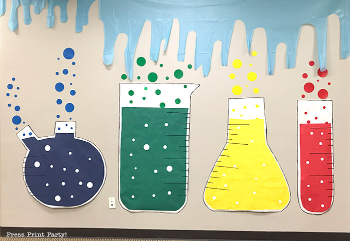 4 giant paper beakers for science party decorations wall backdrop. red, blue, yellow and green with bubbles and slime coming down the wall.