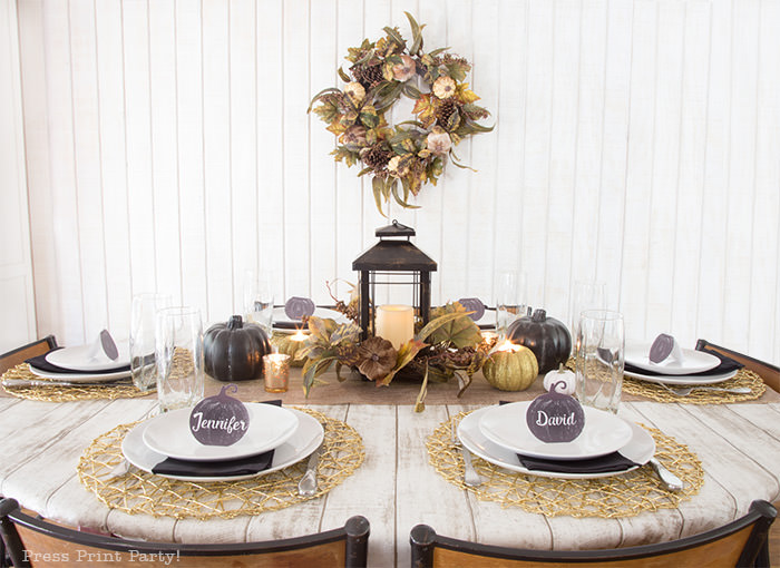 Golden Farmhouse Decorations for Thanksgiving Table. Get inspired with Thanksgiving table decor ideas. White wooden walls with gilded wreath and gold chargers, white plates, black pumpkin place cards, black lantern, golden pumpkins, votives, decorations for thanksgiving table, farmhouse decor ideas, Friendsgiving ideas, thanksgiving table decor, Thanksgiving table setting - tablescape. Press Print Party!