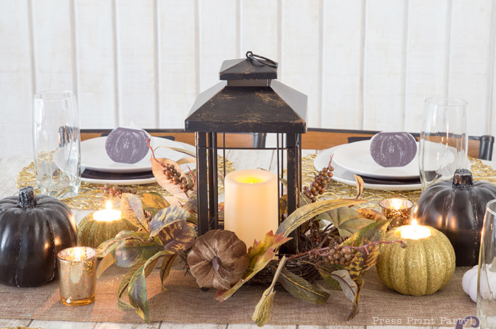 Golden Farmhouse Decorations for Thanksgiving Table. Get inspired with Thanksgiving table decor ideas. White wooden walls with gilded wreath and gold chargers, white plates, black pumpkin place cards, black lantern, golden pumpkins, votives, decorations for thanksgiving table, farmhouse decor ideas, Friendsgiving ideas, thanksgiving table decor, Thanksgiving table setting - tablescape. Press Print Party!