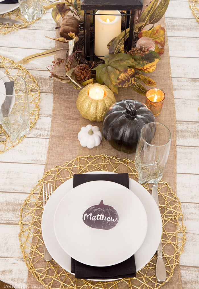 Golden Farmhouse Decorations for Thanksgiving Table. Get inspired with Thanksgiving table decor ideas. White wooden walls with gilded wreath and gold chargers, white plates, black pumpkin place cards, black lantern, golden pumpkins, votives, decorations for thanksgiving table, farmhouse decor ideas, Friendsgiving ideas, thanksgiving table decor, Thanksgiving table setting - tablescape. Press Print Party!