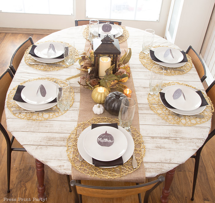 Golden Farmhouse Decorations for Thanksgiving Table. Get inspired with Thanksgiving table decor ideas. White wooden walls with gilded wreath and gold chargers, white plates, black pumpkin place cards, black lantern, golden pumpkins, votives, decorations for thanksgiving table, farmhouse decor ideas, Friendsgiving ideas, thanksgiving table decor, Thanksgiving table setting - tablescape. Press Print Party!