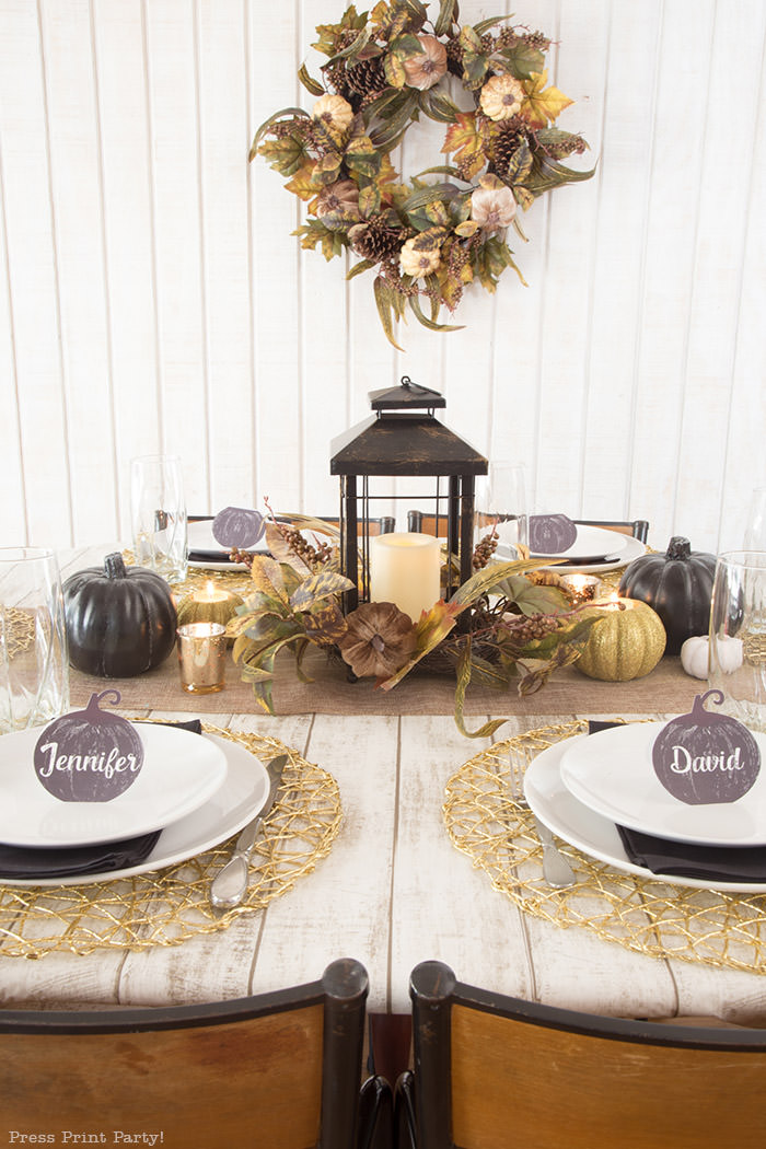 Golden Farmhouse Decorations for Thanksgiving Table. Get inspired with Thanksgiving table decor ideas. White wooden walls with gilded wreath and gold chargers, white plates, black pumpkin place cards, black lantern, golden pumpkins, votives, decorations for thanksgiving table, farmhouse decor ideas, Friendsgiving ideas, thanksgiving table decor, Thanksgiving table setting - tablescape. Press Print Party!