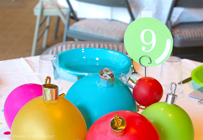 close up of large ornament christmas centerpiece with ballons. with table number 9. Balloons with ornament toppers in green, gold, blue, red and hot pink. Christmas tablescape balloon centerpiece decoration with large ornament balloons and plates that look like ornaments. Very cheery and bright. Fun and creative for your Christmas party. Simple Christmas balloon decor ideas diy. Press Print Party!