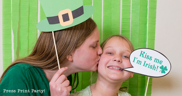 St. Patrick's day free photo booth props printables. girl with Leprechaun hat kissing girl with kiss me I'm Irish sign by Press Print Party!