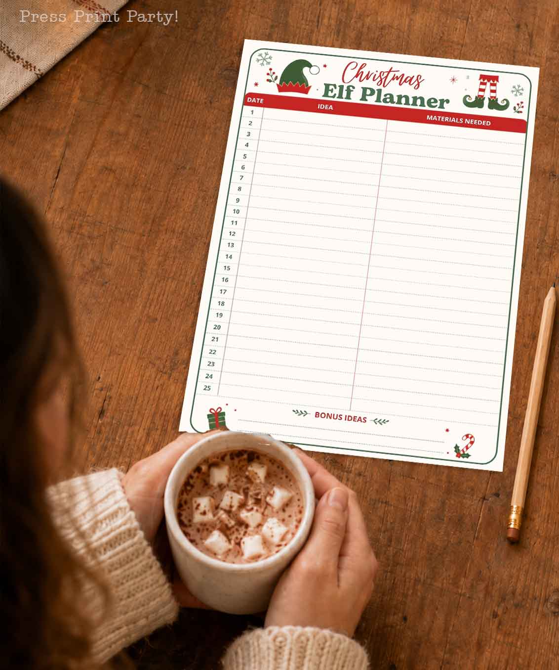 A person holding a mug of hot chocolate with marshmallows sits at a wooden table beside an elf on the shelf printable Christmas Elf Planner sheet and a pencil.