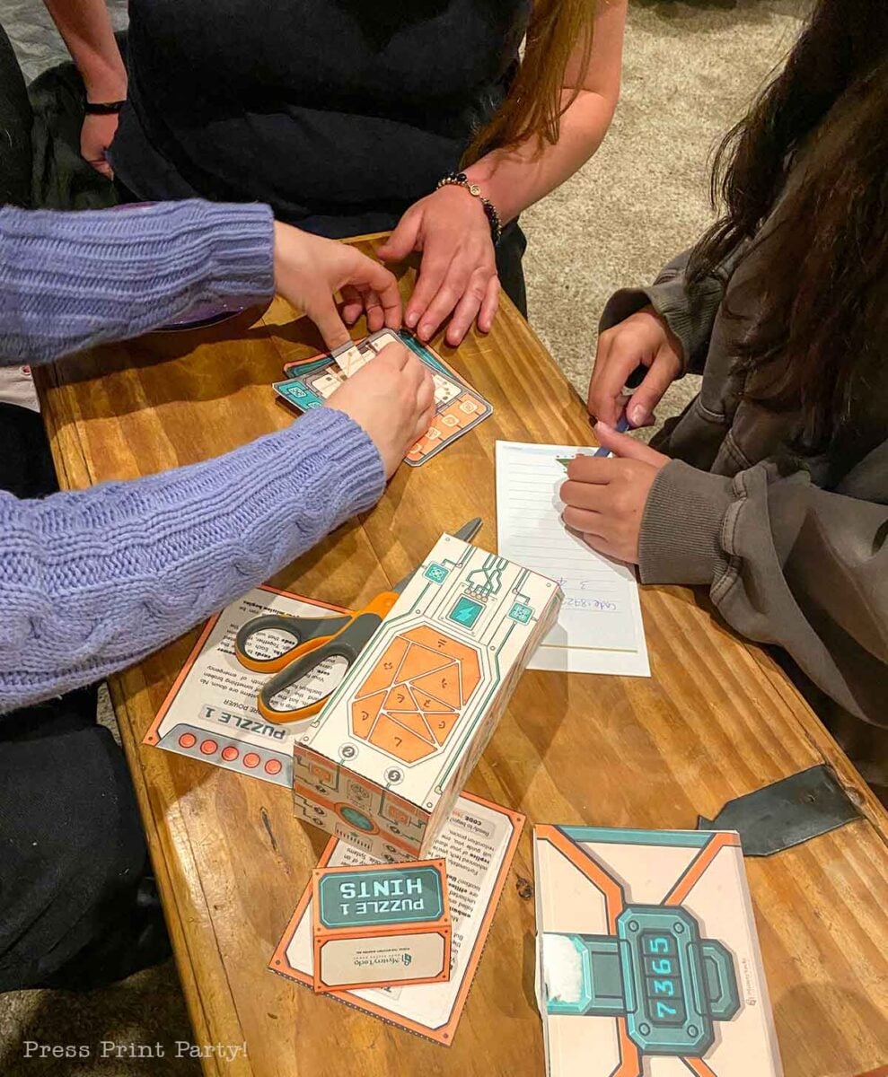 Three people interact with escape room puzzle cards and clues on a wooden table, making it an engaging option for teen birthday party ideas. One person writes on a notepad while the others handle the puzzle pieces. Scissors, hint cards, and a “Puzzle Hints” box are visible, adding to the fun and collaborative atmosphere perfect for celebrating special occasions.