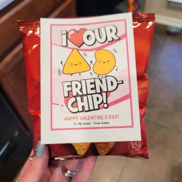 A hand holds a "Kids Valentine Chip Bag Tag Pink, I ❤ Our Friend-chip" Doritos bag featuring a card with happy chip and nacho stating, "I love our friend-chip! Happy Valentine’s Day! To: my friend From: Kellan.