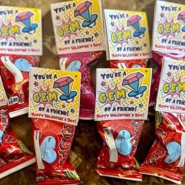 Several "Kids Valentine Chip Bag Tag Pink" packages, featuring the message "I ❤ Our Friend-chip," are arranged with attached Valentine's Day cards, showcasing colorful gem illustrations against a textured brown surface, evoking the delight of finding surprises in chip bags.