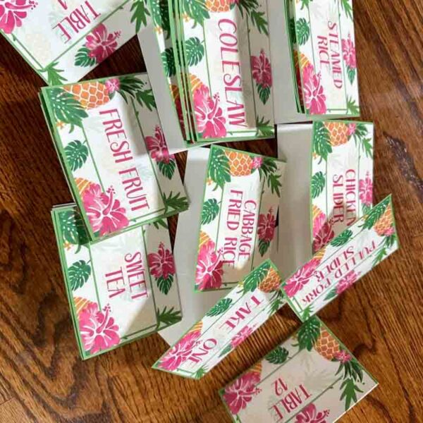 Tropical-themed place cards on a wooden surface highlight fresh fruit, cabbage fried rice, and sweet tea. Decorated with pink flowers and green leaves, they exude vibrant summer vibes—ideal when paired with the "Kids Valentine Chip Bag Tag Pink" for a relaxed feast.