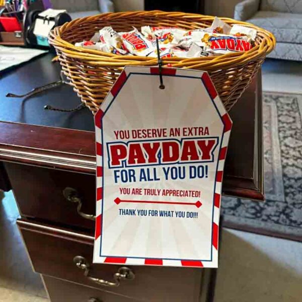 A basket with PayDay candy bars and a colorful Kids Valentine Chip Bag Tag Pink is on a wooden desk. A sign reads, "You deserve an extra PayDay for all you do! You are truly appreciated! Thank you!.