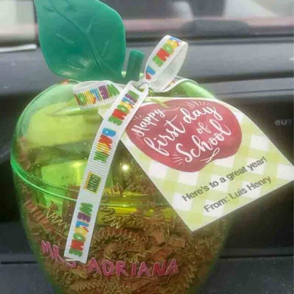 A chip bag-shaped, green apple container on a car dashboard is filled with gold paper shreds. It has a "Happy first day of school" tag and a "Welcome" ribbon, with the name "Mrs. Adriana" on it. The style resembles the "Kids Valentine Chip Bag Tag Pink, I ❤ Our Friend-chip.