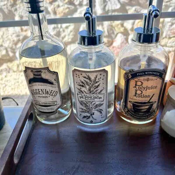 Three glass bottles with pump dispensers sit on a tray, labeled as potions: Wiggenweld, Pepperup, and Polyjuice. Their vintage-style labels are reminiscent of the Kids Valentine Chip Bag Tag Pink, I ❤ Our Friend-chip, adorned with botanical designs and backlit by sunlight through a window.