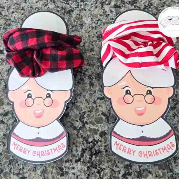 On a granite surface, two cartoon Grandma cutouts with "Merry Christmas" text hold twisted hairbands, one resembling a red and white striped chip bag and another in red and black plaid.