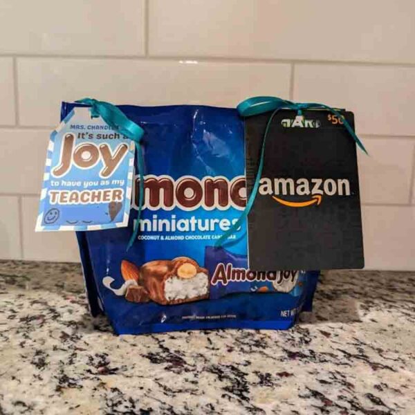 A gift package sits on a granite countertop, featuring Almond Joy miniatures, a chip bag with the tag "I ❤ Our Friend-chip," and an Amazon gift card. The bags are elegantly adorned with teal ribbons. A tag reads, "It's such a joy to have you as my teacher.
