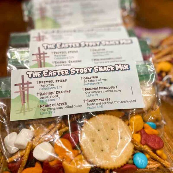 Packets labeled "Kids Valentine Chip Bag Tag Pink, I ❤ Our Friend-chip" resemble classic chip bags and contain snacks like pretzel sticks, raisins, marshmallows, and crackers with biblical references. These packets are neatly stacked on a wooden surface.