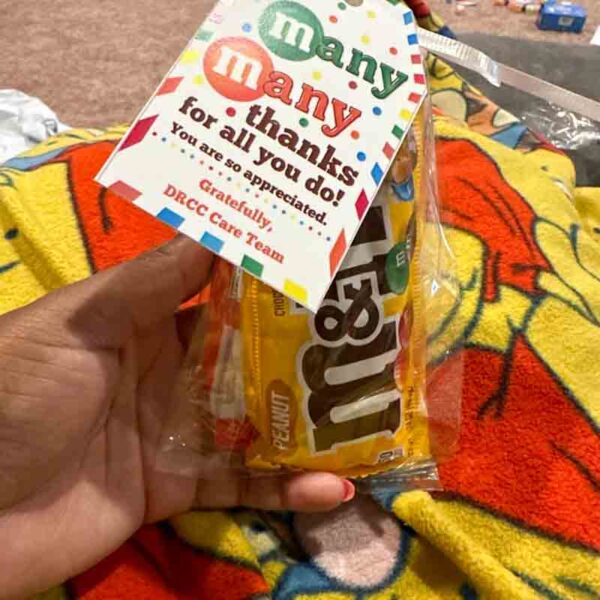 A hand holds a Kids Valentine Chip Bag Tag Pink, "I ❤ Our Friend-chip" on a bag of peanut M&M's, reading "Many thanks for all you do! You are so appreciated. Gratefully, DRCC Care Team." Against a colorful blanket backdrop, it exudes the warmth of sharing with friends.