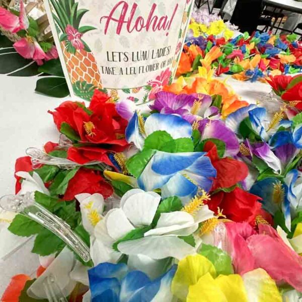 A vibrant array of Hawaiian leis with red, blue, white, yellow, and pink flowers is set on a table next to an "I ❤ Our Friend-chip" chip bag. A festive "Aloha!" sign invites ladies to take a lei or two.