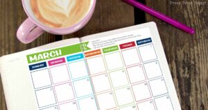 A blank March 2025 calendar for bullet journaling is spread open on a wooden table, featuring colorful borders for each day. A pink coffee mug with latte art and a purple pen sit stylishly beside the calendar.