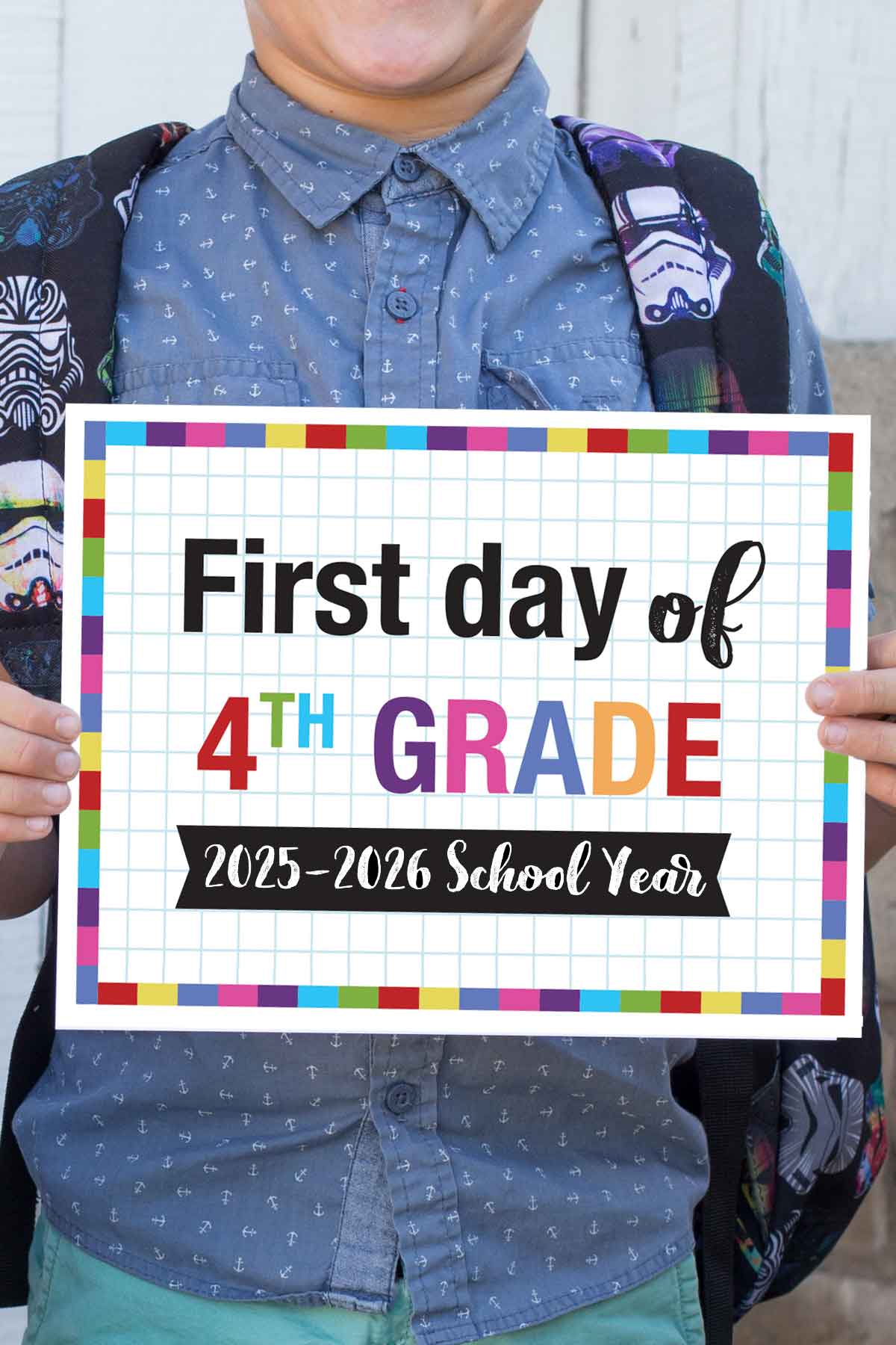 A child wearing a blue patterned shirt holds a colorful first day of school printable sign that says "First day of 4th grade, 2025-2026 school year" with a grid background and multicolored border.