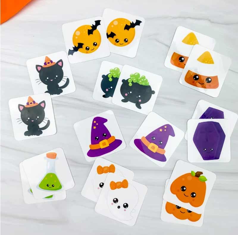 Colorful Halloween-themed matching game cards featuring cute illustrations, including a black cat, candy corn, witch's hat, bubbling cauldron, ghost, pumpkin, and moon with bats, spread out on a white marble surface.