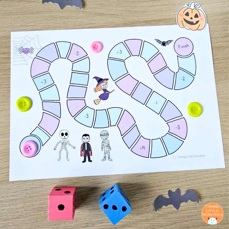 Colorful Halloween-themed board game with a winding path featuring numbered squares, spooky illustrations like a witch, skeleton, vampire, mummy, spider, and bat. The game board is surrounded by buttons used as player markers and two wooden dice, all set on a wooden table.