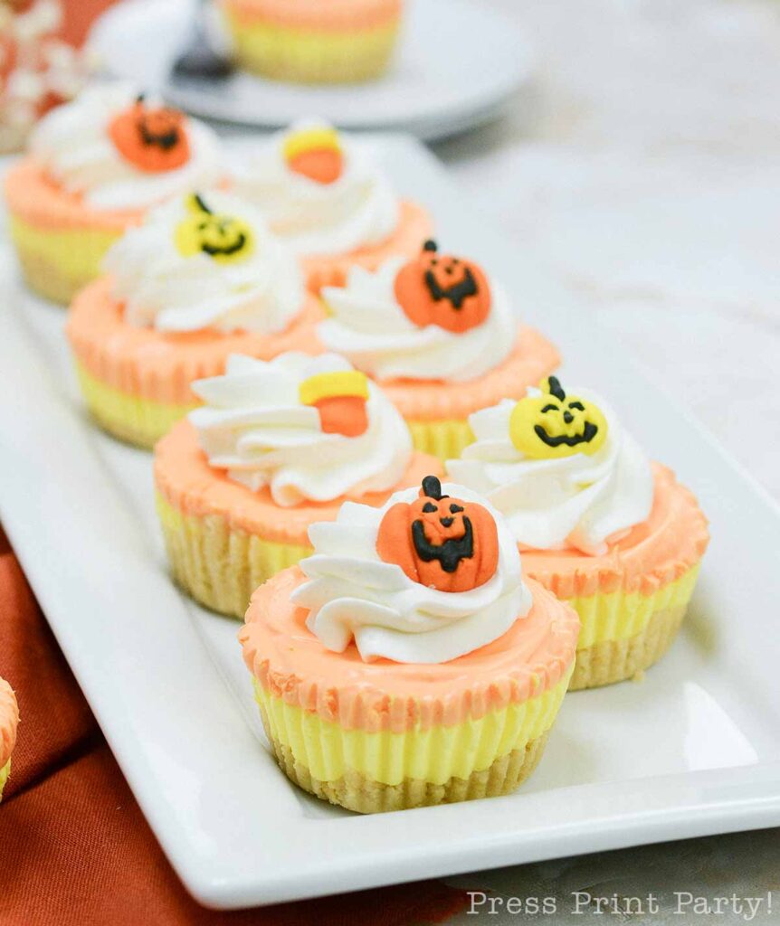 A rectangular white plate holds several mini candy corn cheesecakes topped with swirls of whipped cream and small Halloween-themed decorations, including jack-o'-lantern faces. The cheesecakes have an orange outer layer and a creamy center. A blurred background shows more desserts.