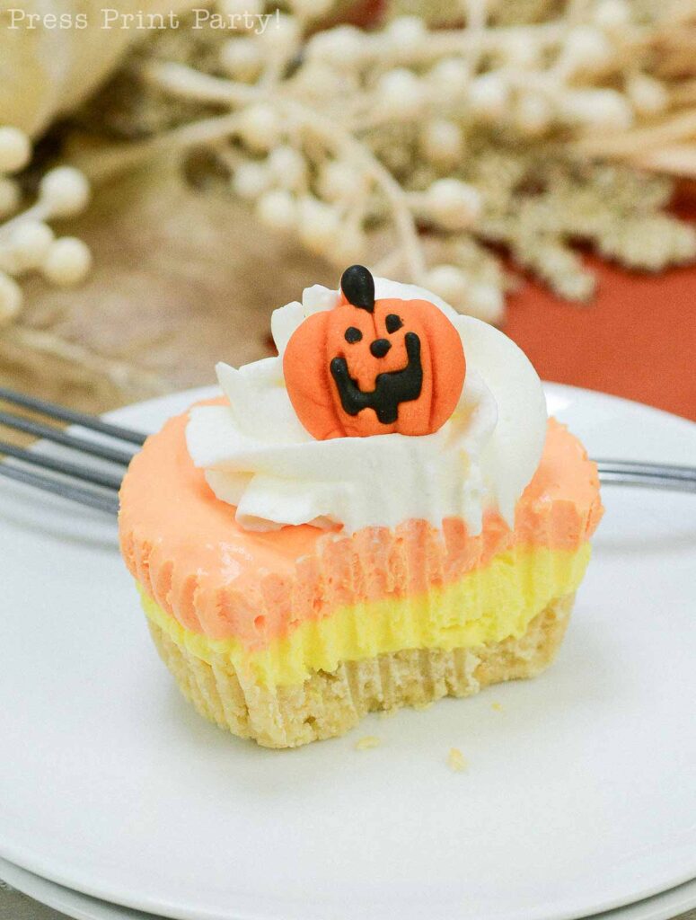 A Halloween-themed candy corn cheesecake cupcake with layers of orange and yellow frosting, topped with white whipped cream and a pumpkin decoration with a smiling face. The cupcake is placed on a white plate with a fork nearby. Background shows festive fall decor.