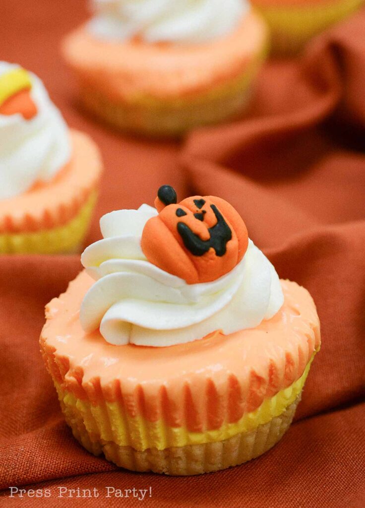 A close-up of a decorated cupcake with orange icing and white swirls, topped with a small pumpkin face candy. The cupcake is set on an orange fabric background, resembling the festive appeal of candy corn cheesecake, with more similarly decorated cupcakes in the blurred background.