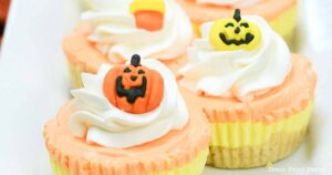 A close-up view of Halloween-themed cupcakes topped with swirls of orange and white frosting. Each cupcake, decorated with orange pumpkin faces showing varied expressions, sits neatly arranged on a white plate beside a delectable slice of candy corn cheesecake.