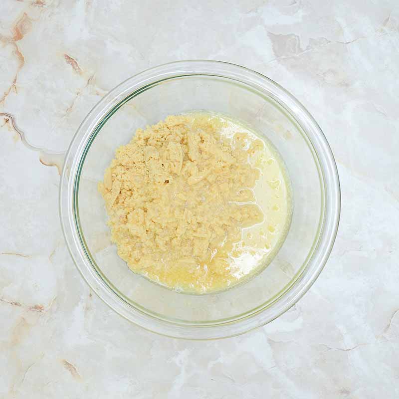 A clear glass bowl on a marbled countertop containing a mixture of dry and wet ingredients for candy corn cheesecake. The mixture appears to be partially mixed, with chunks of dry batter and pools of liquid visible.