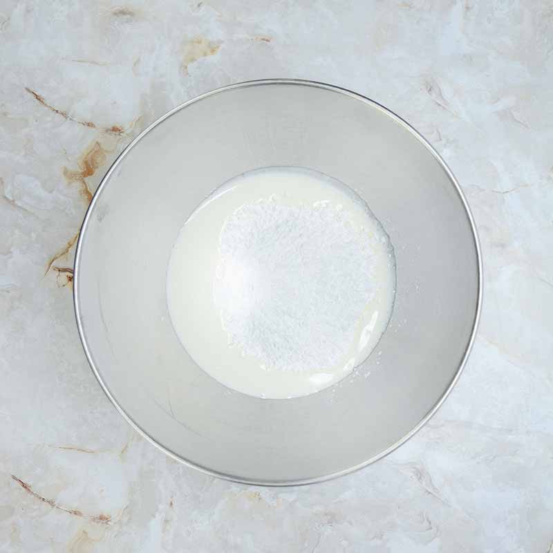 A stainless steel mixing bowl containing a small amount of white powder, likely sugar or flour, combined with liquid, possibly milk or cream, sits on a light marbled surface. The ingredients appear to be in the early stages of mixing for a candy corn cheesecake recipe.