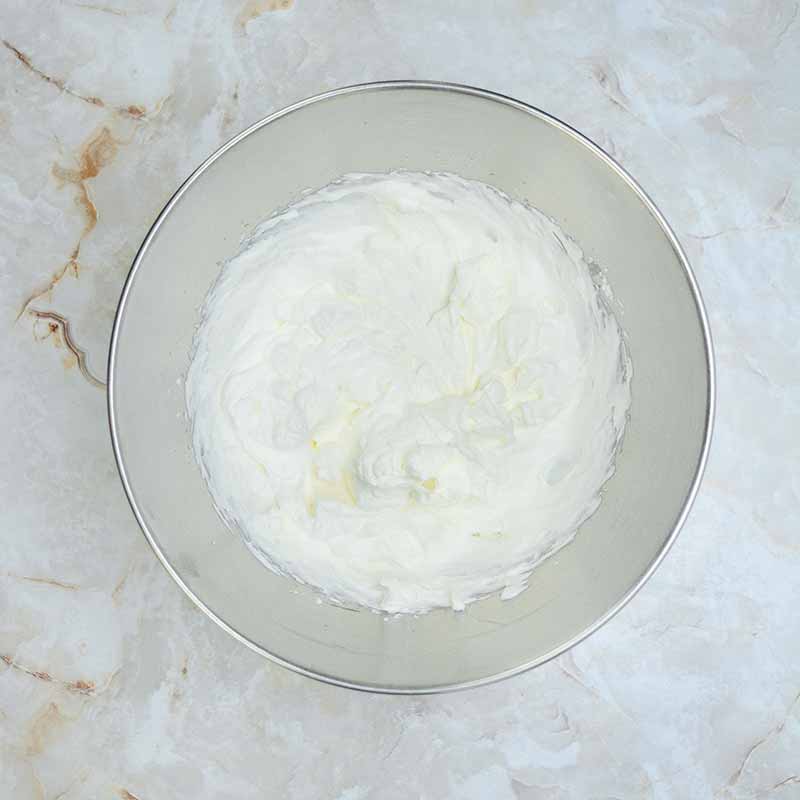 A stainless steel mixing bowl filled with whipped cream sits on a marbled countertop. The whipped cream has a light, fluffy texture and slightly uneven surface, perfect for topping a candy corn cheesecake.