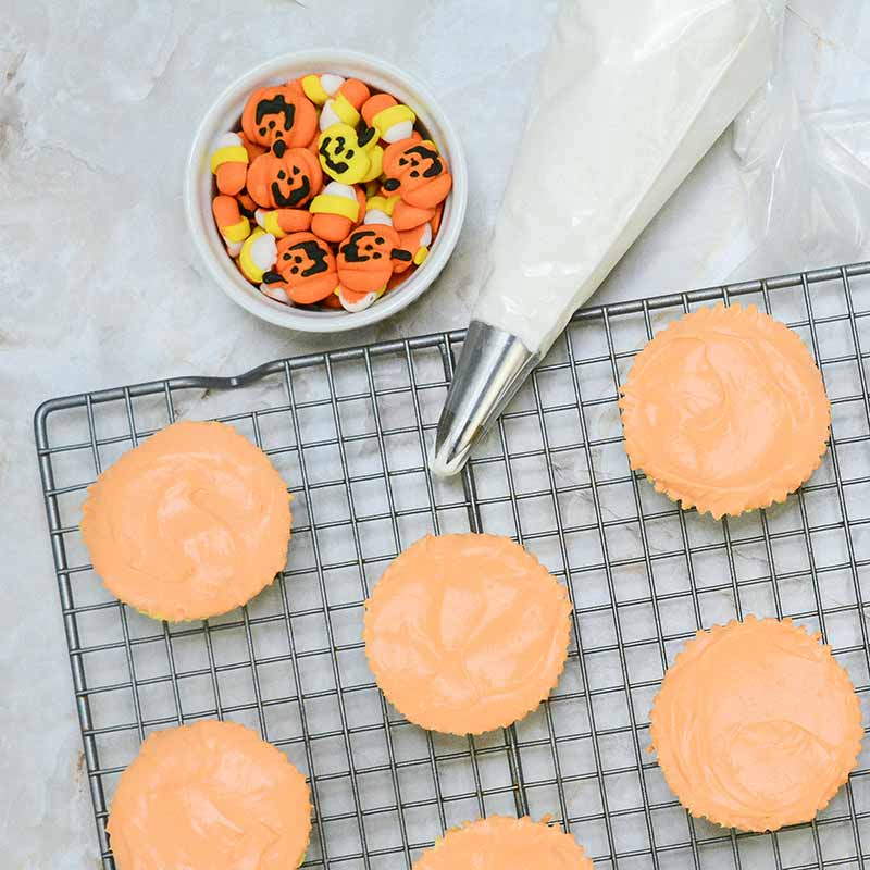 A wire cooling rack with six cupcakes topped with orange frosting sits on a marble surface. To the upper left, there's a small bowl filled with Halloween-themed candy, including pieces decorated with pumpkin faces and candy corn cheesecake bites. A piping bag filled with frosting is nearby.