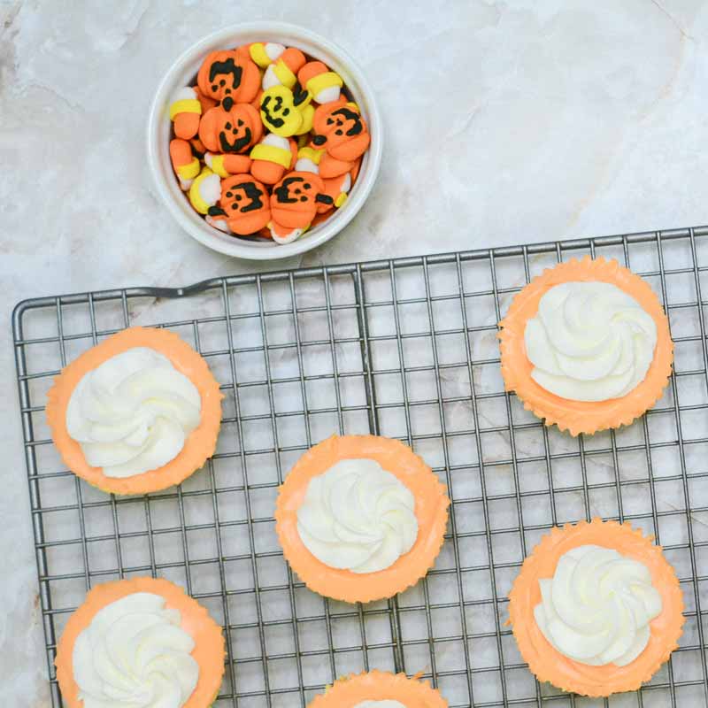 A cooling rack holds six orange and white frosted cupcakes. Nearby, a white bowl filled with Halloween-themed candy, including small jack-o'-lantern faces and candy corn cheesecake bites, sits on a light-colored marble surface.
