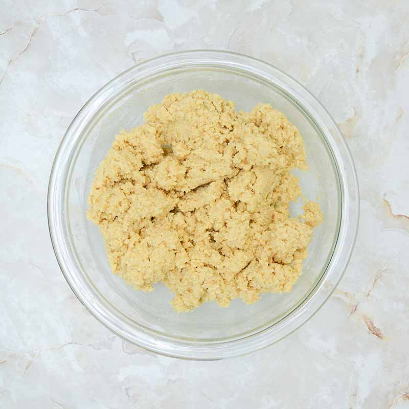 A glass bowl filled with light brown, crumbly cookie dough that hints at a delicious candy corn cheesecake sits on a marbled countertop. The dough appears to be mixed but not yet formed into individual cookies.