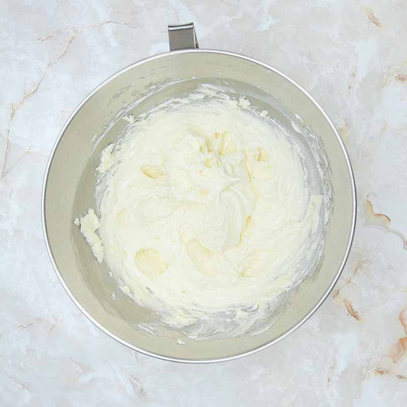 A stainless steel mixing bowl containing smooth, creamy whipped butter, reminiscent of candy corn cheesecake, set on a white marble countertop. The whipped butter has a slightly fluffy texture with some soft peaks.
