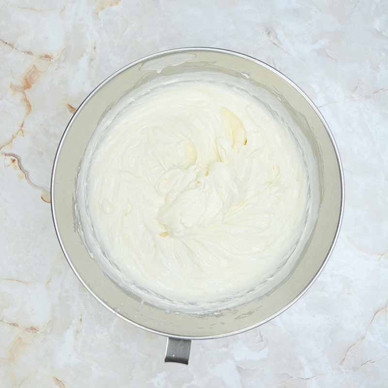 A metallic bowl containing creamy, smooth, white candy corn cheesecake batter sits on a marble surface. The mixture looks well-whipped and has a glossy texture.
