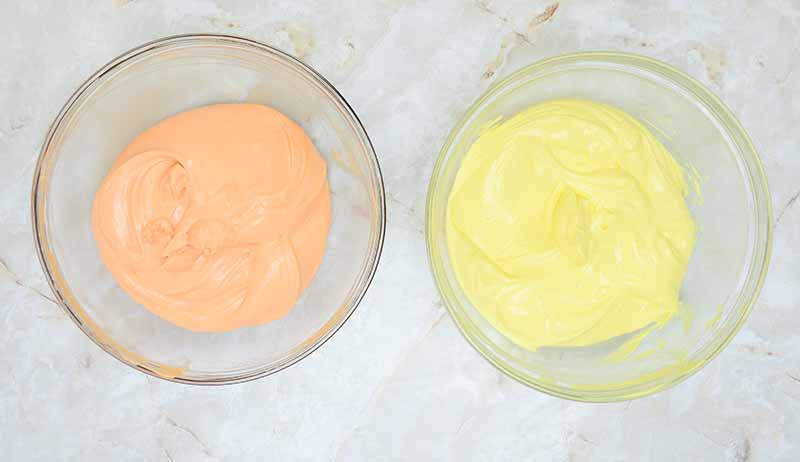Two glass bowls sit on a light-colored, marble countertop. One bowl contains smooth, orange-colored frosting or batter, and the other holds a similar smooth mixture in yellow. The vibrant hues evoke the festive spirit of a candy corn cheesecake, perfectly placed side by side.