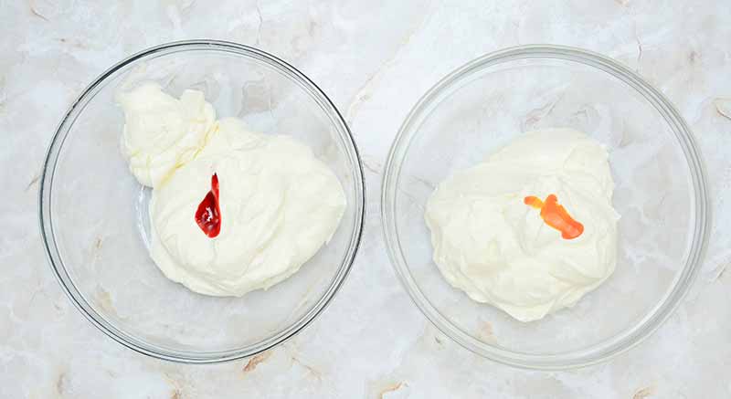Two clear glass bowls placed on a marble surface are filled with creamy white frosting. The bowl on the left has a small dollop of red food coloring, while the bowl on the right has a small dollop of orange food coloring, giving them the look of candy corn cheesecake.